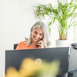 Lächelnde Person nimmt ein Telefonat entgegen, hinter einem Monitor sitzend.