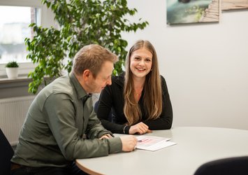 Zwei Personen am Tisch, eine junge Frau schaut in die Kamera, ein Mann erklärt eine ausgedruckte Liste.