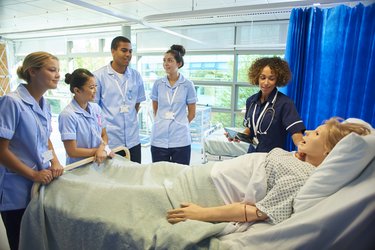 Vier Auszubildende im Skills Lab stehen gemeinsam mit ihrer Lehrkraft am Krankenbett, in dem eine Pflegepuppe liegt, die unterschiedliche Krankheiten und Reaktionen simulieren kann